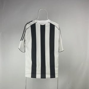 Pu&ograve; includere: Una maglia sportiva a righe bianche e nere, probabilmente per una squadra di calcio, &egrave; appesa a un gancio. La maglia ha un girocollo e maniche corte con bordi neri. Il retro della maglia &egrave; visibile.