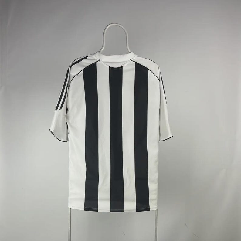 Pu&ograve; includere: Una maglia sportiva a righe bianche e nere, probabilmente per una squadra di calcio, &egrave; appesa a un gancio. La maglia ha un girocollo e maniche corte con bordi neri. Il retro della maglia &egrave; visibile.