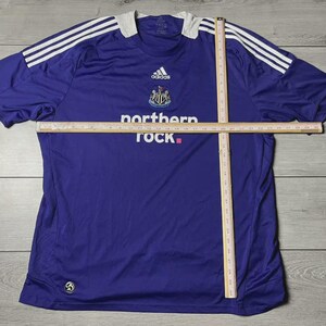 May include: A purple Newcastle United football shirt by Adidas, with white shoulder stripes and a white collar. It displays the club crest, Adidas logo, and the 'northern rock' sponsor logo.