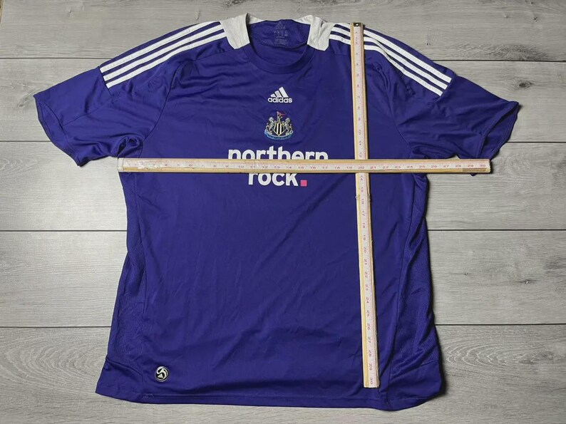 May include: A purple Newcastle United football shirt by Adidas, with white shoulder stripes and a white collar. It displays the club crest, Adidas logo, and the 'northern rock' sponsor logo.