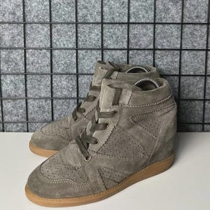 May include: A pair of gray suede high-top sneakers with a wedge heel. The sneakers have a perforated design on the sides, brown laces, and a brown rubber sole. The shoes are displayed on a white surface.