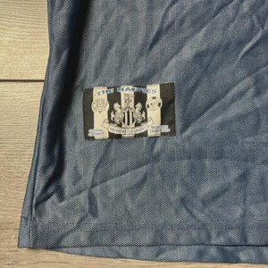 K&ouml;nnte beinhalten: Nahaufnahme eines blauen Fu&szlig;balltrikots mit strukturiertem Muster. Ein rechteckiger Aufn&auml;her zeigt "THE MAGPIES" &uuml;ber einem Wappen und "NEWCASTLE UNITED". Das Trikot besteht aus leichtem, atmungsaktivem Stoff.