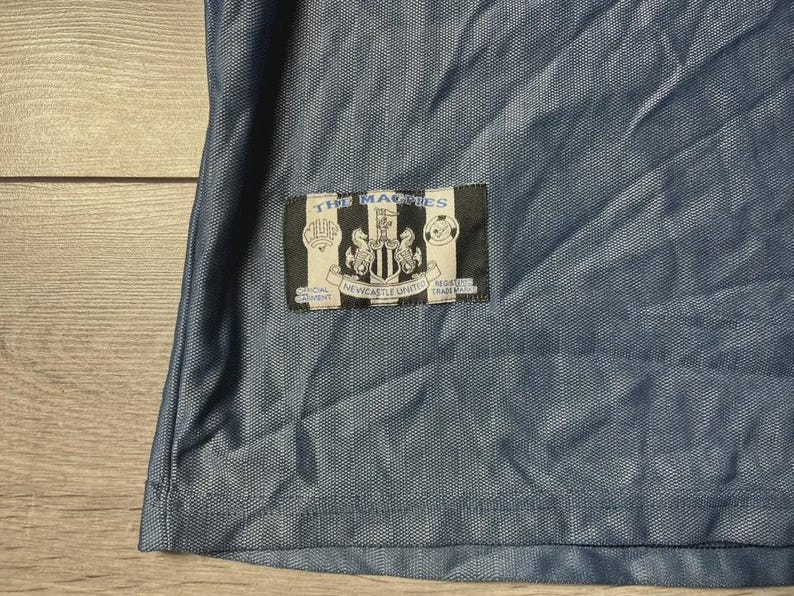 K&ouml;nnte beinhalten: Nahaufnahme eines blauen Fu&szlig;balltrikots mit strukturiertem Muster. Ein rechteckiger Aufn&auml;her zeigt "THE MAGPIES" &uuml;ber einem Wappen und "NEWCASTLE UNITED". Das Trikot besteht aus leichtem, atmungsaktivem Stoff.