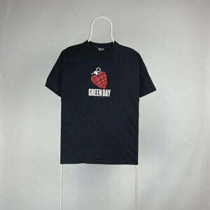 May include: Black t-shirt featuring a red heart-shaped grenade graphic with the text "GREEN DAY" in white. The shirt is displayed on a white hanger against a neutral background. A classic band tee.