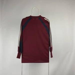 May include: Maroon long-sleeve athletic shirt with navy blue and white accents. The shirt features white stripes down the sleeves and navy blue panels on the shoulders. The shirt is hanging on a white hanger.