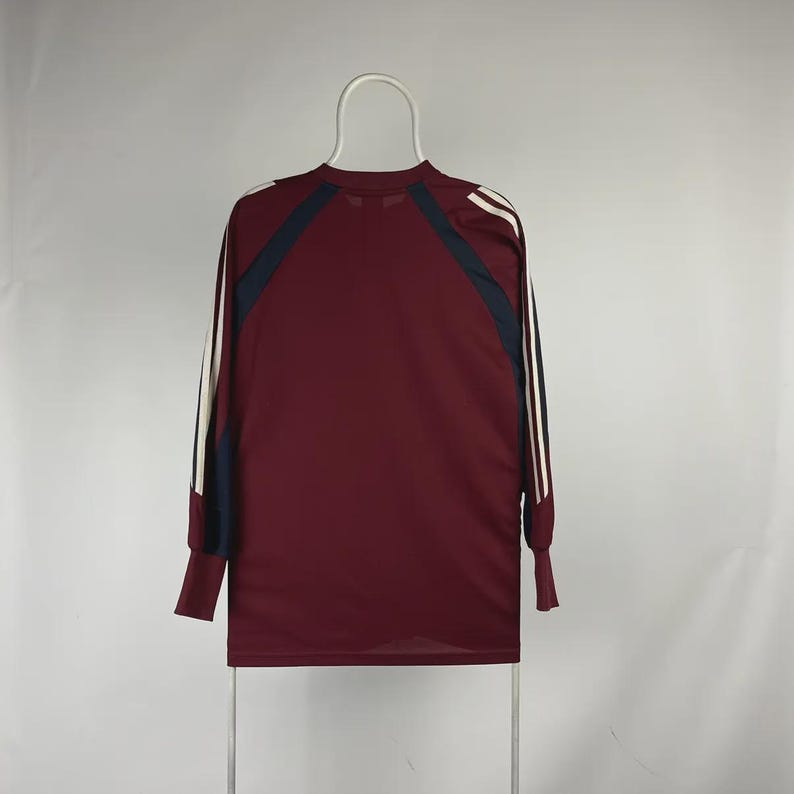 May include: Maroon long-sleeve athletic shirt with navy blue and white accents. The shirt features white stripes down the sleeves and navy blue panels on the shoulders. The shirt is hanging on a white hanger.