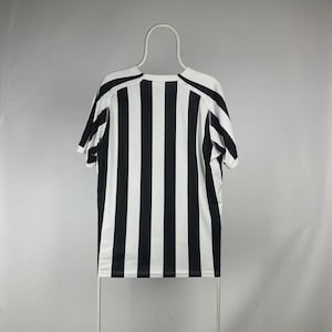 Puede incluir: Una camiseta de manga corta a rayas blancas y negras colgada de una percha blanca. La camiseta tiene rayas verticales y cuello redondo. Las mangas y los hombros tienen un dise&ntilde;o negro liso.