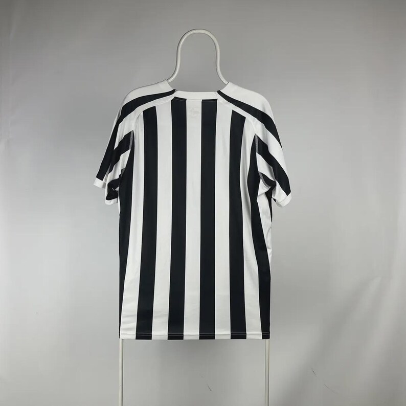 Puede incluir: Una camiseta de manga corta a rayas blancas y negras colgada de una percha blanca. La camiseta tiene rayas verticales y cuello redondo. Las mangas y los hombros tienen un dise&ntilde;o negro liso.
