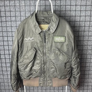 May include: Olive green bomber jacket with a zip closure, two front pockets, and a ribbed hem. Features the text "Tannkosh" and a patch with a logo. The jacket is hanging on a white hanger against a grey grid background.