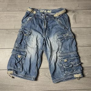以下が含まれることがあります： 複数のポケットとベージュのベルトが付いた、青いデニムカーゴショーツ。 ショーツは色あせたウォッシュ加工で、木製の表面に平らに置かれています。 ウエストバンドには「JUST LAG」の文字が見えます。
