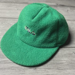 Könnte beinhalten: Eine leuchtend grüne Baseballkappe aus Fleece mit gebogenem Schirm. Auf der Kappe befindet sich ein gestickter Text in Lila, Blau und Weiß auf der Vorderseite. Der Hut besteht aus einem weichen, strukturierten Material.
