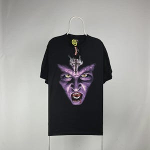 May include: Black t-shirt featuring a purple and gray graphic of a vampire face with fangs, yellow eyes, and a cross. The shirt is hanging on a white hanger against a plain background. The design is centered on the front of the shirt.