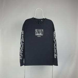 May include: Black long-sleeved shirt with a white Keith Haring graphic on the front and white line art on the sleeves. The graphic on the front shows a person with a turntable. The shirt is hanging on a white hanger.