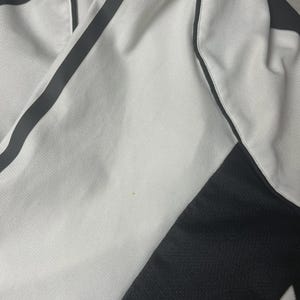 Pu&ograve; includere: Primo piano di un capo di abbigliamento sportivo bianco con dettagli neri. Il tessuto ha un aspetto strutturato, con una striscia nera sul lato. Il capo sembra essere realizzato in un materiale traspirante.