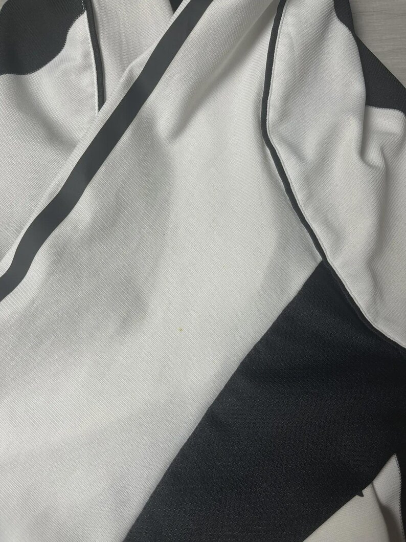 Pu&ograve; includere: Primo piano di un capo di abbigliamento sportivo bianco con dettagli neri. Il tessuto ha un aspetto strutturato, con una striscia nera sul lato. Il capo sembra essere realizzato in un materiale traspirante.