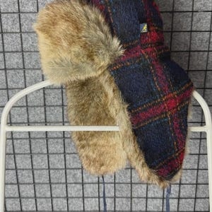 Peut inclure: Un chapeau d'hiver avec un motif à carreaux bleu marine et rouge, orné d'une garniture en fausse fourrure marron. Le chapeau a des cache-oreilles et une attache bleue. Un petit logo jaune est visible sur le côté.