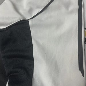 Pu&ograve; includere: Primo piano di una maglia sportiva bianca e nera. La maglia presenta un corpo bianco con dettagli neri sui lati e un pannello nero sulla spalla. Una striscia grigia corre lungo il lato. Il tessuto ha un aspetto strutturato.