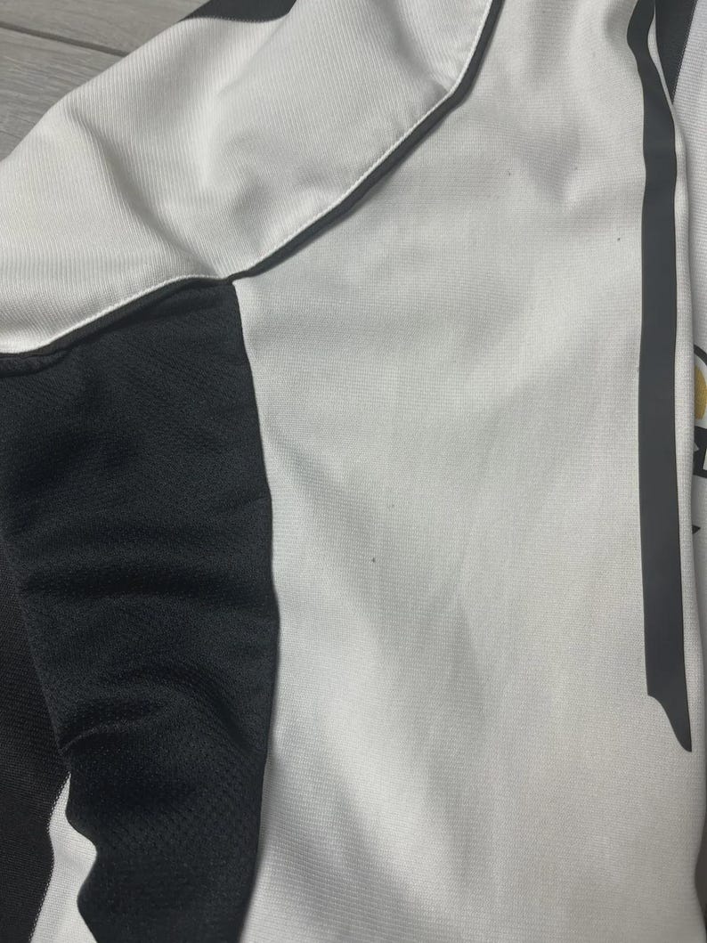 Pu&ograve; includere: Primo piano di una maglia sportiva bianca e nera. La maglia presenta un corpo bianco con dettagli neri sui lati e un pannello nero sulla spalla. Una striscia grigia corre lungo il lato. Il tessuto ha un aspetto strutturato.