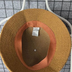 Puede incluir: Un sombrero de pescador marr&oacute;n con un exterior texturizado. El interior presenta una banda color melocot&oacute;n y una etiqueta blanca. El sombrero se muestra en una percha blanca sobre un fondo de rejilla gris. Este accesorio es elegante.