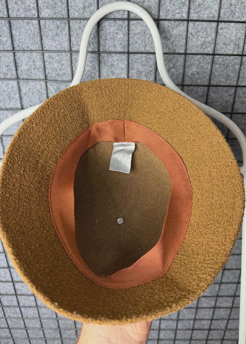 Puede incluir: Un sombrero de pescador marr&oacute;n con un exterior texturizado. El interior presenta una banda color melocot&oacute;n y una etiqueta blanca. El sombrero se muestra en una percha blanca sobre un fondo de rejilla gris. Este accesorio es elegante.