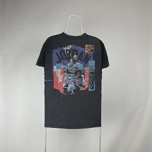 May include: Black t-shirt featuring a graphic of a basketball player with the word "JORDAN" in blue and red. The number "23" is visible on the player's jersey. A Nike logo is in the upper right corner. The shirt is hanging on a white hanger.