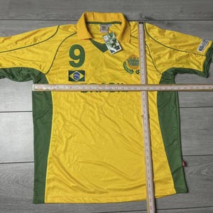 May include: Yellow and green Brazil soccer jersey with the number 9 on the front. The jersey has a collar and short sleeves. The Brazilian flag is on the front. A ruler is placed across the jersey.