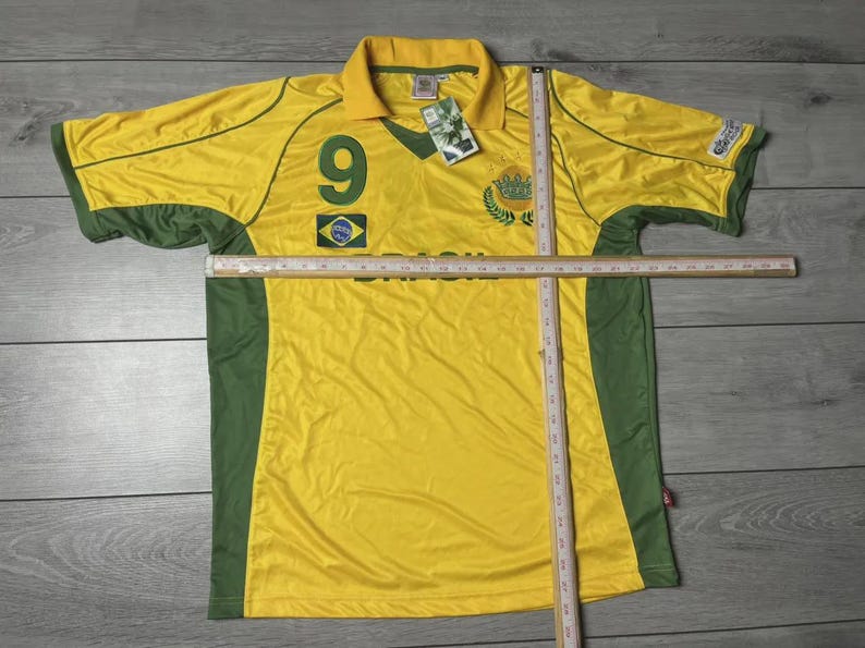 May include: Yellow and green Brazil soccer jersey with the number 9 on the front. The jersey has a collar and short sleeves. The Brazilian flag is on the front. A ruler is placed across the jersey.