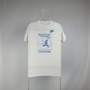 May include: White t-shirt featuring a blue graphic for the Belfast City Marathon 1982, with the Nike logo and Guinness branding. The shirt is hanging on a white hanger against a neutral background.