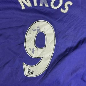 May include: A close-up of a purple jersey. The name "NIKOS" is printed in white across the top. Below, a large white number "9" is visible, outlined in black, with a small emblem at the bottom. The jersey is made of a textured fabric.