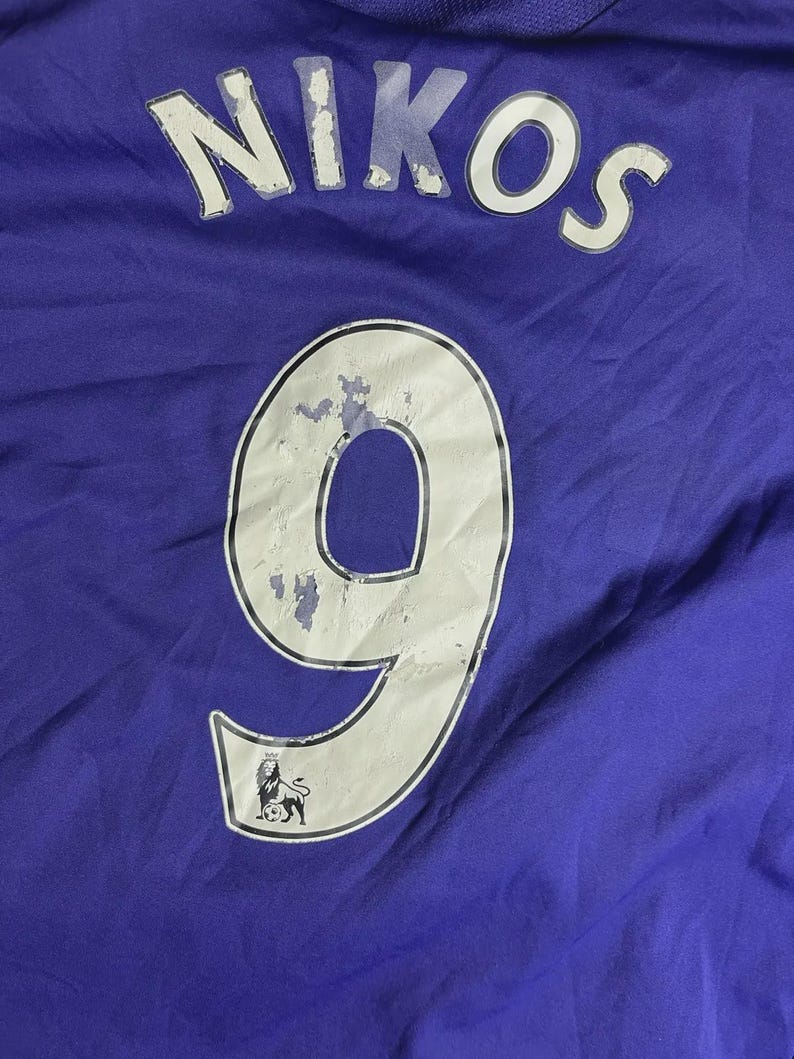 May include: A close-up of a purple jersey. The name "NIKOS" is printed in white across the top. Below, a large white number "9" is visible, outlined in black, with a small emblem at the bottom. The jersey is made of a textured fabric.