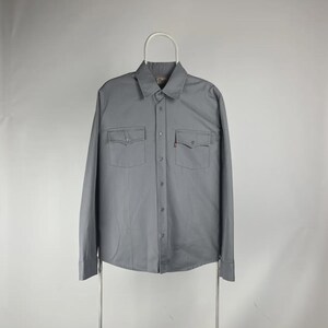 May include: A long-sleeve button-down shirt in a solid light gray color. The shirt features a standard collar, a central button placket, and two chest pockets with buttoned flaps. A small red tab is visible on the edge of the left chest pocket.