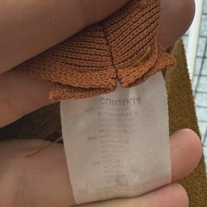 Puede incluir: Primer plano de una tela de punto marr&oacute;n con una etiqueta blanca. La etiqueta dice "CONTENTS" y detalla la composici&oacute;n de la tela, incluyendo acr&iacute;lico. La tela tiene un aspecto texturizado.