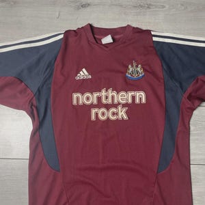 May include: Maroon and navy long-sleeve soccer jersey with the text "northern rock" in white. Features the Adidas logo and a team crest. White stripes accent the navy sleeves.