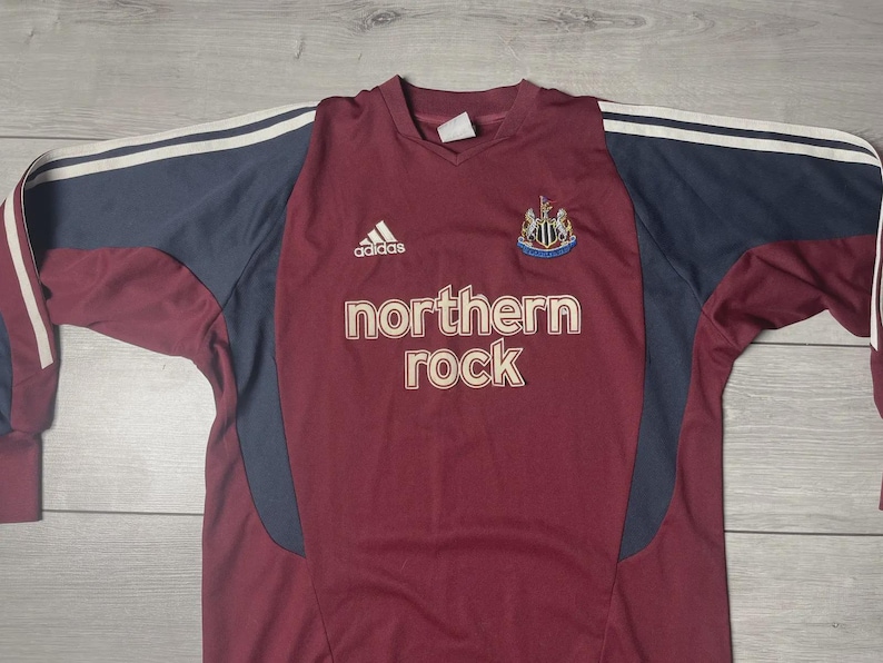 May include: Maroon and navy long-sleeve soccer jersey with the text "northern rock" in white. Features the Adidas logo and a team crest. White stripes accent the navy sleeves.