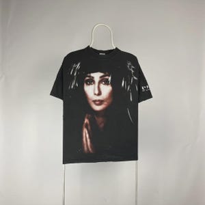 May include: Black t-shirt featuring a portrait of Cher. The image on the shirt is a close-up of Cher's face with dark hair and a feathered headdress. The shirt is hanging on a white hanger.