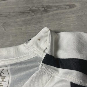Puede incluir: Primer plano de una camiseta deportiva blanca y negra. La camiseta tiene cuello en V y rayas negras en las mangas. El logotipo de Puma es visible en la camiseta. La talla es XL.