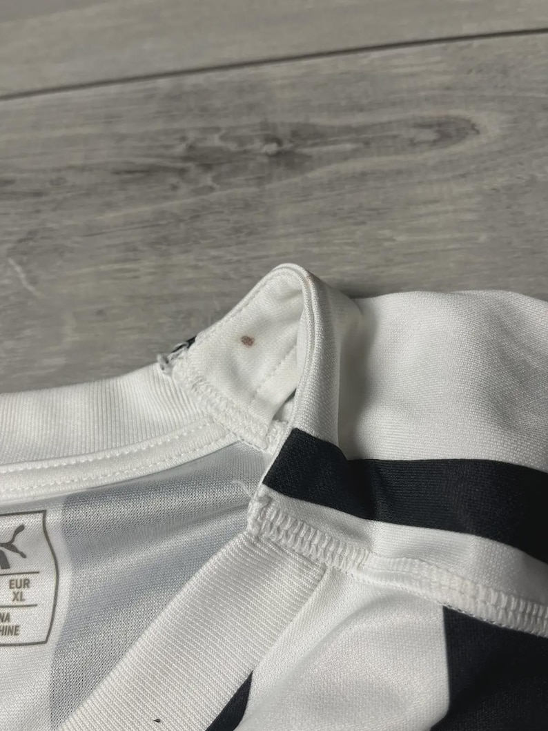 Puede incluir: Primer plano de una camiseta deportiva blanca y negra. La camiseta tiene cuello en V y rayas negras en las mangas. El logotipo de Puma es visible en la camiseta. La talla es XL.