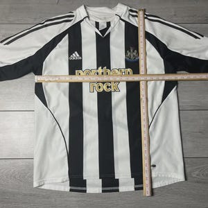 Pu&ograve; includere: Una maglia da calcio a righe bianche e nere con la scritta "northern rock" in oro. La maglia presenta il logo Adidas e lo stemma del Newcastle United. Scollo a V.
