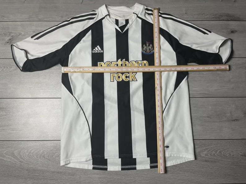 Pu&ograve; includere: Una maglia da calcio a righe bianche e nere con la scritta "northern rock" in oro. La maglia presenta il logo Adidas e lo stemma del Newcastle United. Scollo a V.