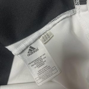 Pu&ograve; includere: Primo piano di una maglia sportiva Adidas a righe bianche e nere. Il logo e il testo Adidas sono visibili su un'etichetta. L'etichetta recita "MADE IN TUNISIA" e "LA MARQUE AUX 3 BANDES".