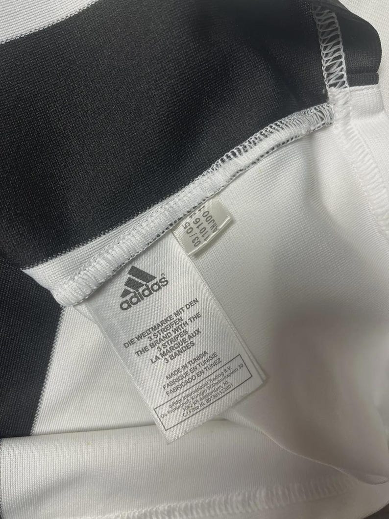 Pu&ograve; includere: Primo piano di una maglia sportiva Adidas a righe bianche e nere. Il logo e il testo Adidas sono visibili su un'etichetta. L'etichetta recita "MADE IN TUNISIA" e "LA MARQUE AUX 3 BANDES".
