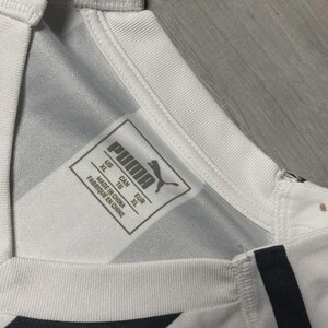 Puede incluir: Primer plano de una camiseta deportiva blanca y negra con el logo de Puma y una etiqueta de talla. La camiseta tiene un patr&oacute;n geom&eacute;trico y un cuello acanalado. La etiqueta indica la talla XL.