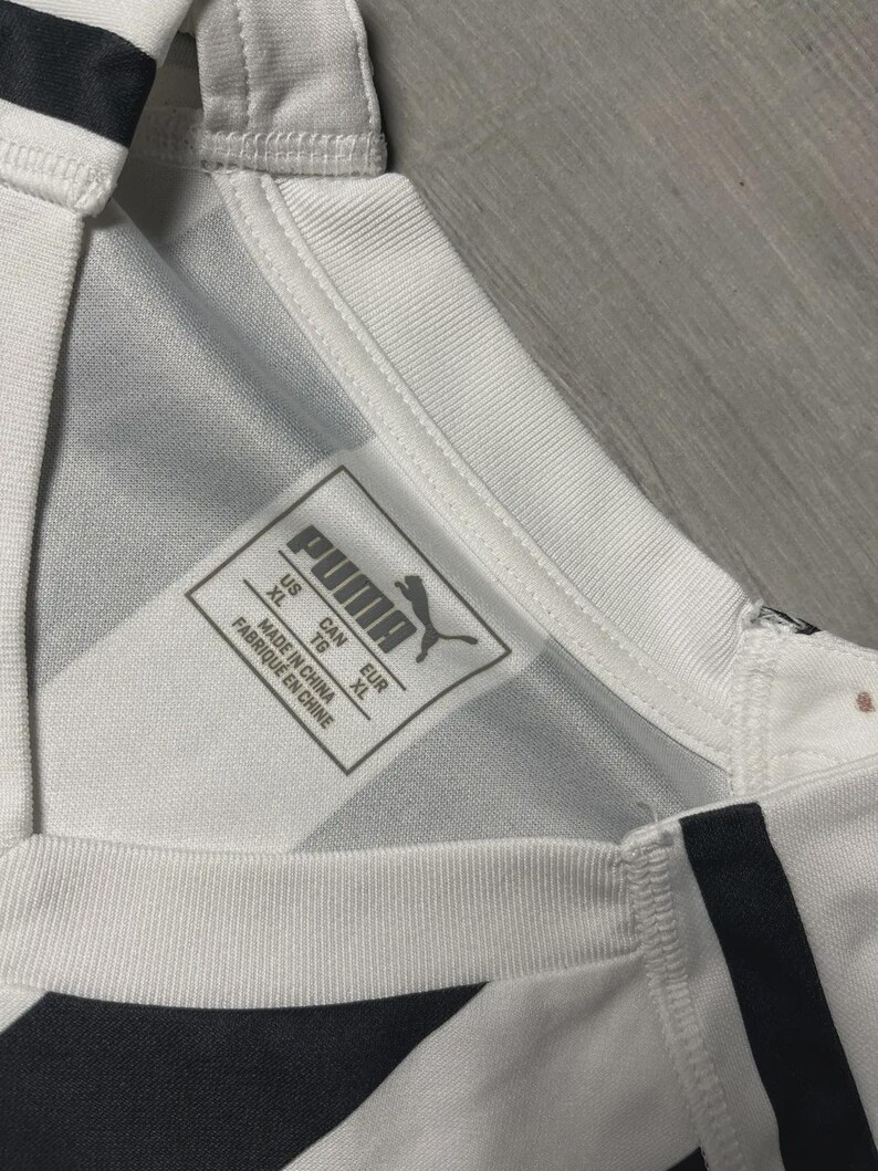Puede incluir: Primer plano de una camiseta deportiva blanca y negra con el logo de Puma y una etiqueta de talla. La camiseta tiene un patr&oacute;n geom&eacute;trico y un cuello acanalado. La etiqueta indica la talla XL.