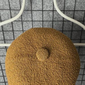 Puede incluir: Boina de color mostaza con un aspecto tejido y texturizado. La boina presenta un peque&ntilde;o detalle redondeado en el centro superior. Se muestra sobre un fondo de rejilla gris y una percha blanca.