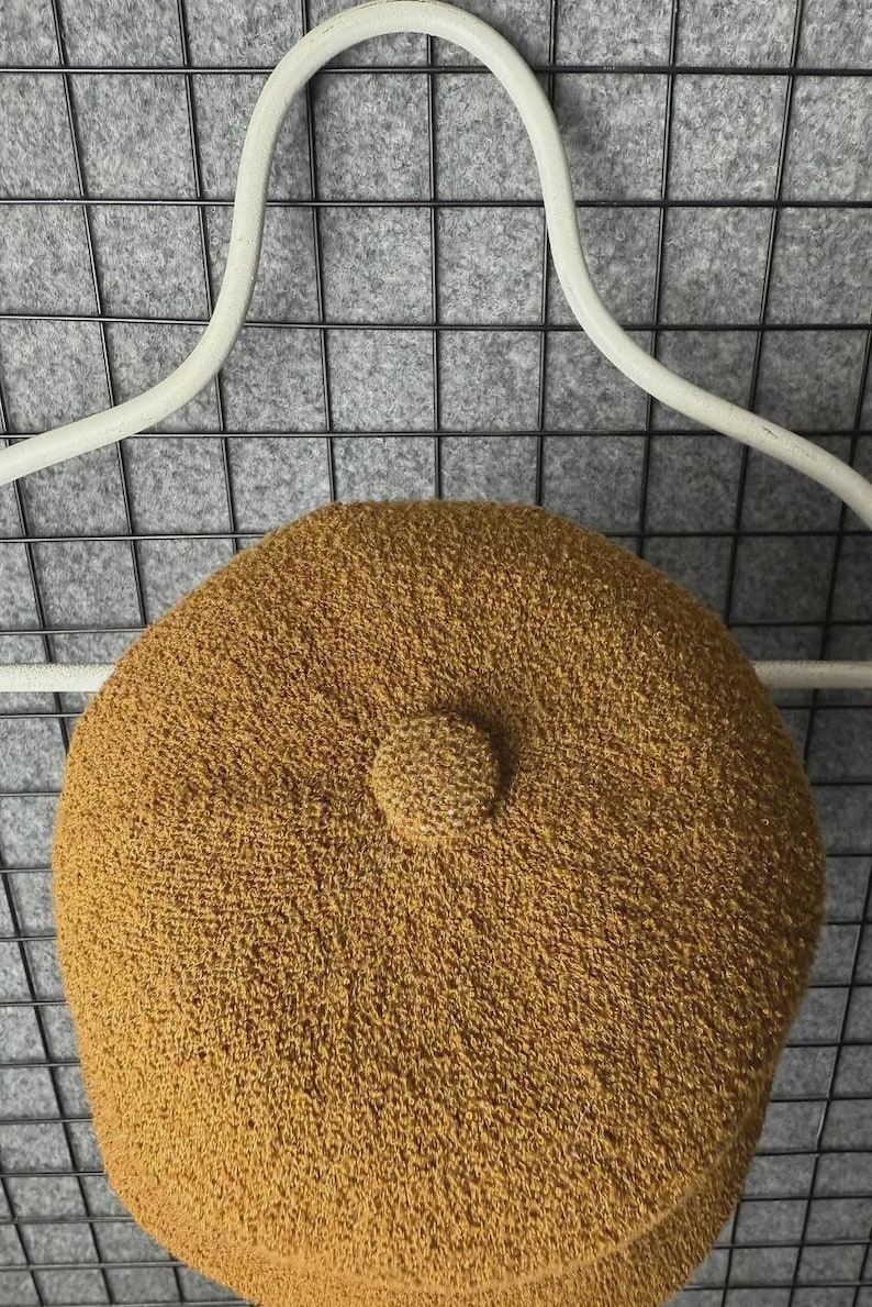 Puede incluir: Boina de color mostaza con un aspecto tejido y texturizado. La boina presenta un peque&ntilde;o detalle redondeado en el centro superior. Se muestra sobre un fondo de rejilla gris y una percha blanca.