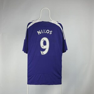 May include: A blue sports jersey with white shoulder and sleeve detailing. The name "NIKOS" and the number "9" are printed in white on the back. The jersey is suspended from a white hanger.