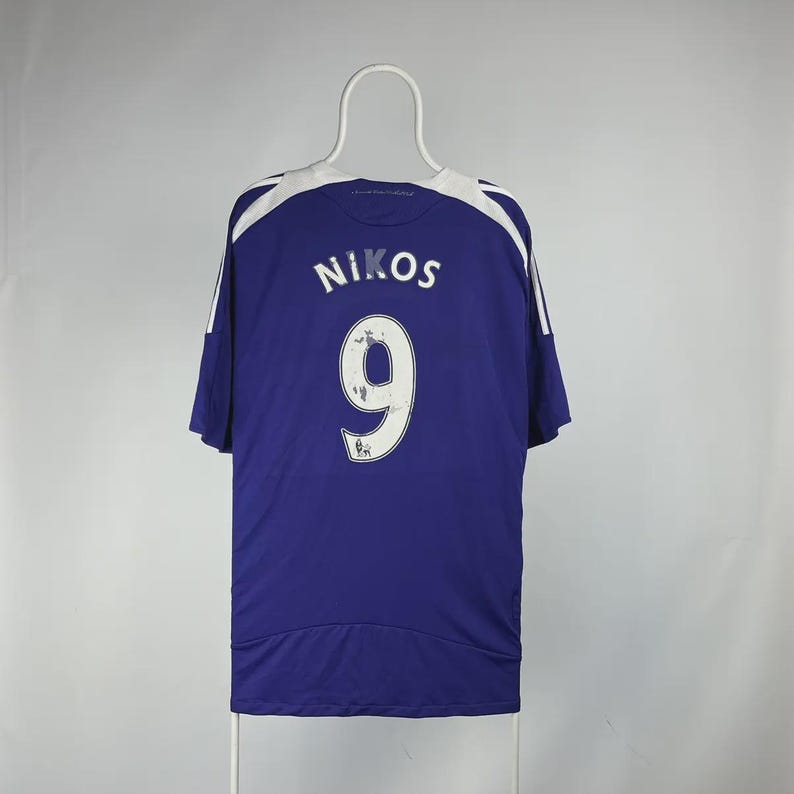May include: A blue sports jersey with white shoulder and sleeve detailing. The name "NIKOS" and the number "9" are printed in white on the back. The jersey is suspended from a white hanger.