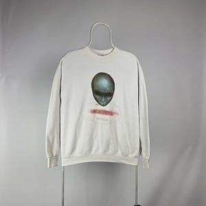 May include: Off-white sweatshirt featuring a large, detailed alien face graphic in shades of gray and green. The graphic includes a red, blurred text. The sweatshirt has a crew neck and long sleeves, displayed on a hanger.