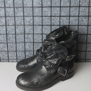 May include: A pair of black leather ankle boots with a distressed, metallic finish. The boots feature buckles, straps, and studded accents. A zipper is visible on the side of one boot. The boots are on a white surface.