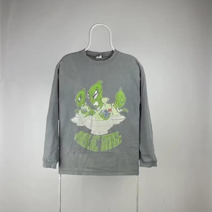 May include: Gray long-sleeve sweatshirt with a graphic of three green aliens on a spaceship. The aliens are holding a globe. The text "PUBLIC IMAGE" is below the graphic in green. The sweatshirt is hanging on a white hanger.